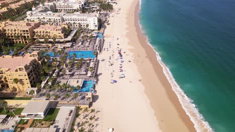 Un-Dron-Aéreo-Vuela-A-Lo-Largo-De-La-Playa-De-Cabo-San-Lucas-Sobre-Las-Piscinas-Al-Aire-Libre-De-Los-Resorts-De-Abajo