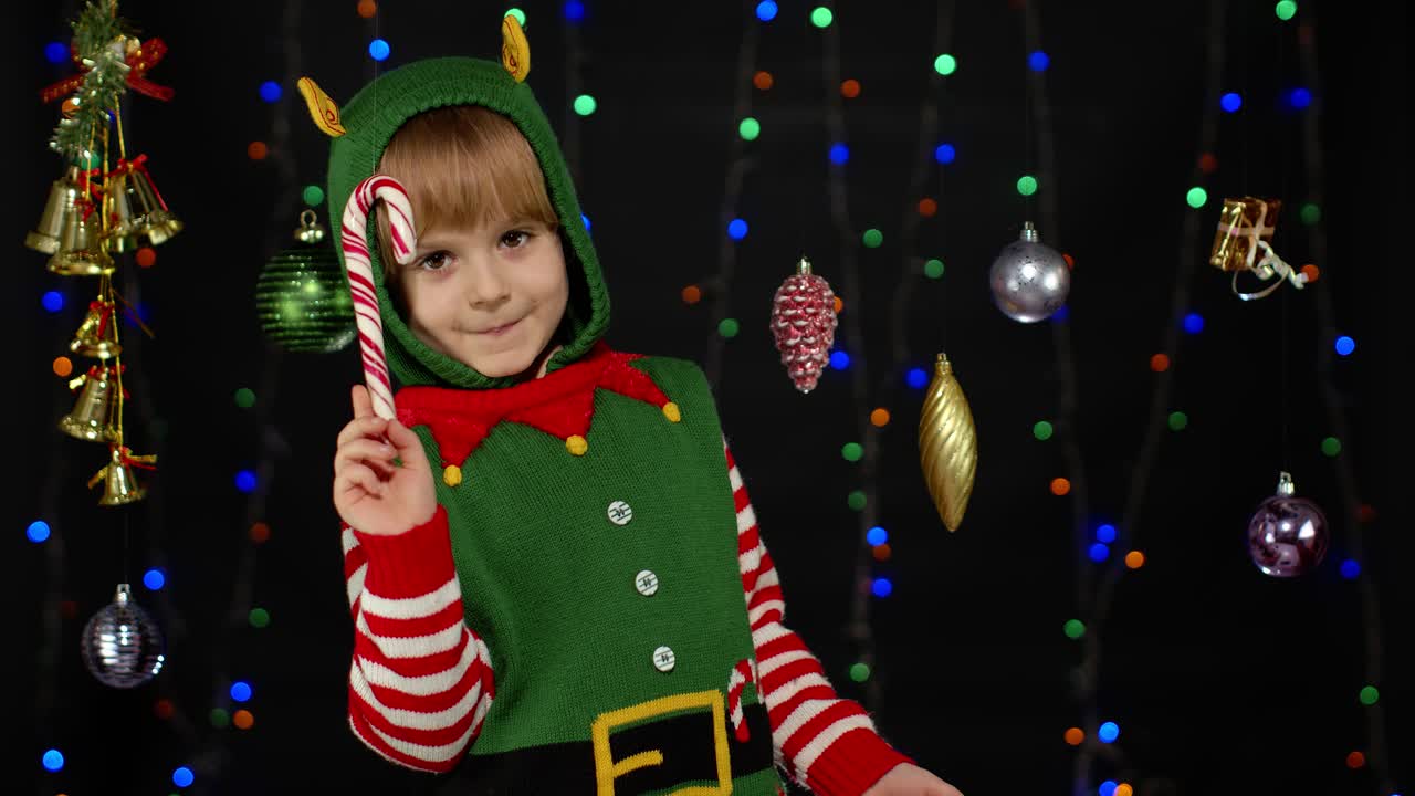 Malá dívka v kostýmu vánočního skřítka pomocníka Santa Clause s lízátkem ve tvaru vánoční hole, radostně se usmívající.