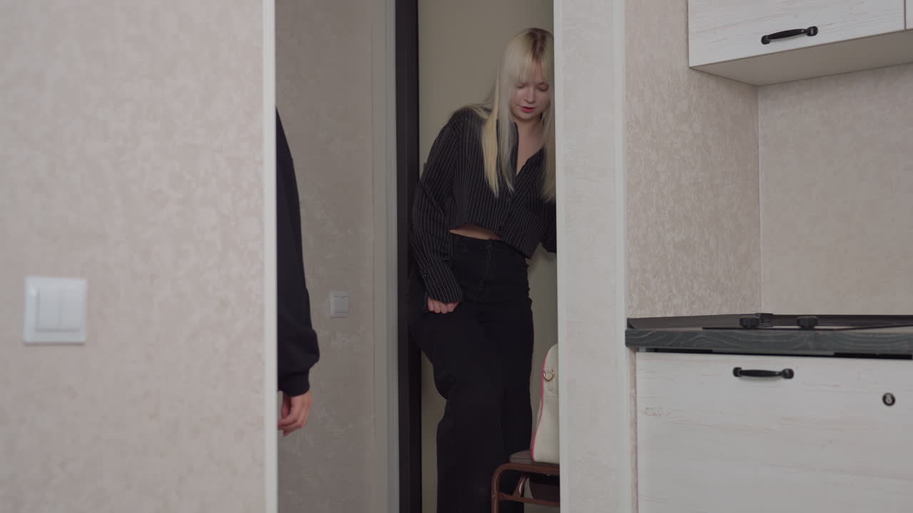 Partial side view of person observing blonde woman in striped shirt slipping on shoe while walking past kitchen area, capturing candid moment of everyday morning routine