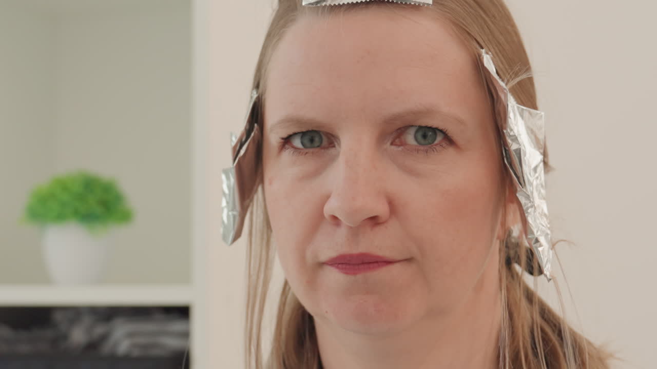 primo piano di una donna in un salone con la stagnola, espressione contemplativa durante la colorazione, mani dell'hair stylist fuori dall'inquadratura, interni moderni e neutri con una pianta in vaso su uno scaffale, mantello protettivo e strisce di stagnola