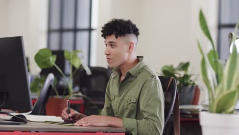 hombre biracial feliz creativo usando tableta y computadora en una oficina casual, en cámara lenta