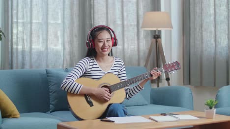 asian woman composer with paper and notebook on table wearing headphones and smiling to camera while playing guitar at home