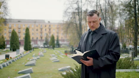 hombre con impermeable negro mirando el horizonte en un cementerio y leyendo una biblia