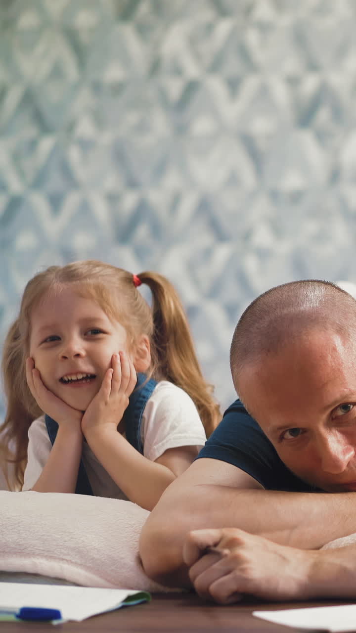 Dolly cam: cheerful little daughter with ponytails and daddy laugh doing homework together on floor with soft blanket in light room at quarantine time