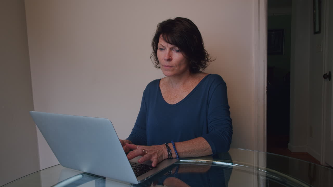 mujer en casa en una computadora portátil escribiendo con expresión facial neutra