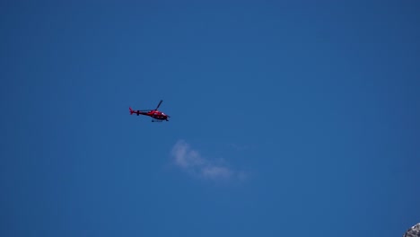 red helicopter next to mountain flying under a blue sky wih clouds