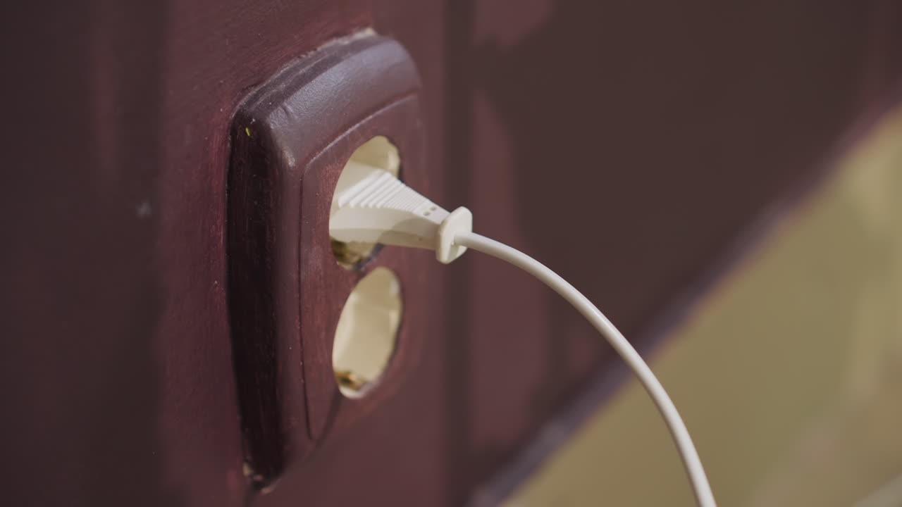 갈색 벽면 콘센트에 손으로 플러그를 꽂는 모습이에요. 흰색 코드를 오래된 콘센트에 천천히, 조심스럽게 연결하는 사람의 클로즈업 샷이며, 따뜻한 실내 톤과 가정적인 분위기가 느껴집니다. 손의 움직임과 정렬에 초점을 맞췄어요.