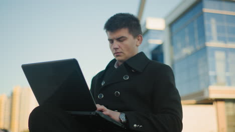 businessman in black coat focused on laptop screen while seated outdoors in urban environment, wristwatch visible on wrist, with modern office building in background