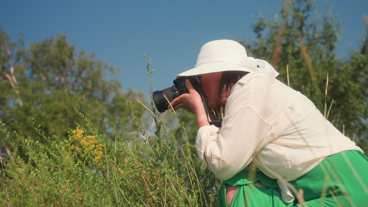 Lady wearing white sun hat and green dress crouches in sunny grassy area focusing camera lens closely on yellow flower while surrounded by tall grass and trees under clear blue sky