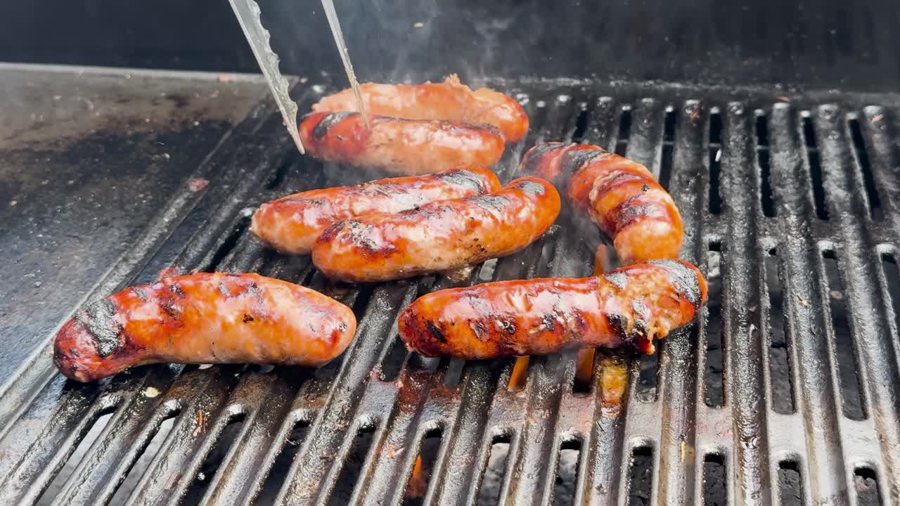 Close-up of sausages being grilled, showing smoke and grill marks. Tongs are used to turn them over.