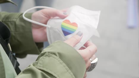 close up of hands with painted nails placing a lgbtq rainbo heart shaped sticker on an ffp2 mask in a city environment on a sunny day