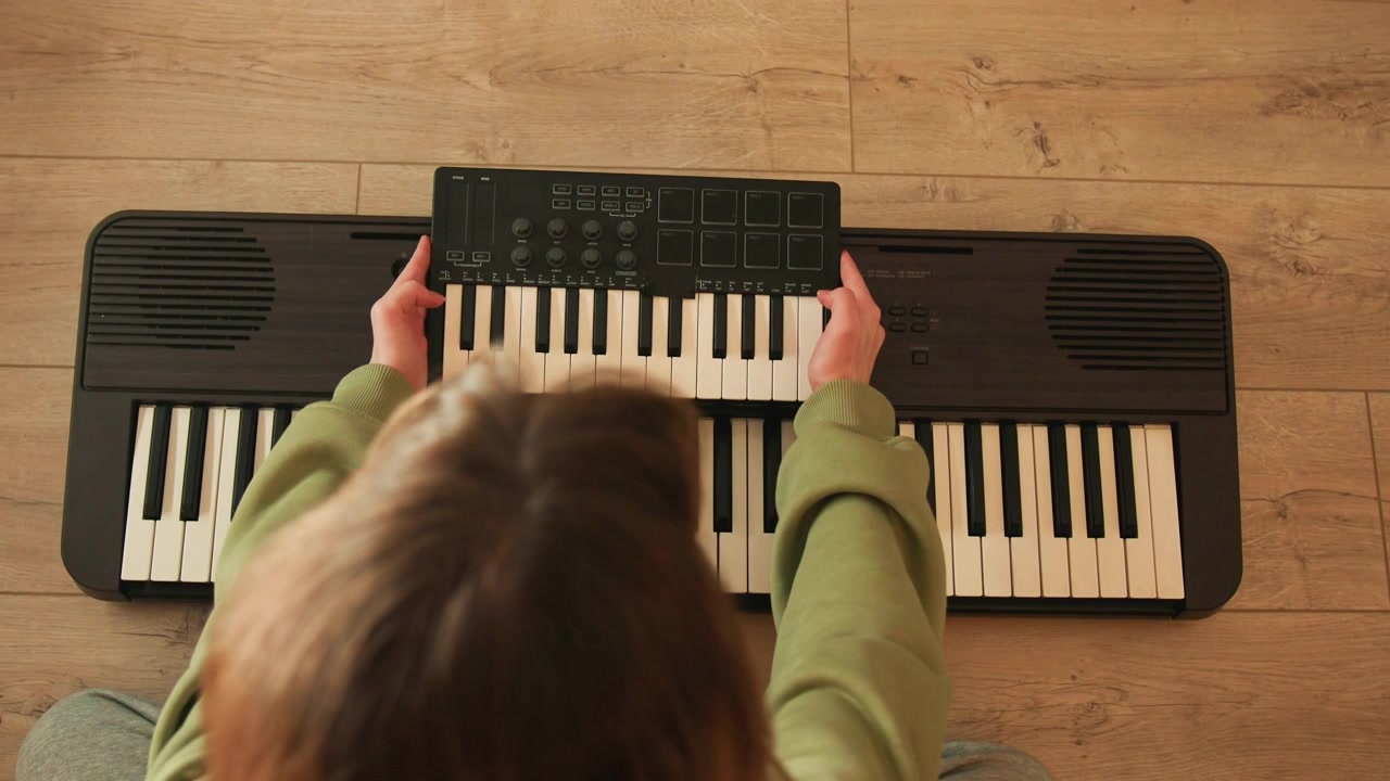 Topdown view of child seated at compact black electronic keyboard, both hands pressing keys across central range while leaning forward in focused posture, showing engaged practice and visible finger