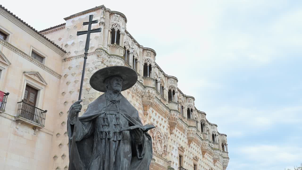 A historic monument statue with a patriarchal cross stands before the Palace of the Dukes of the Infantado in Guadalajara, Spain. It embodies Spanish heritage and striking Renaissance architecture.
