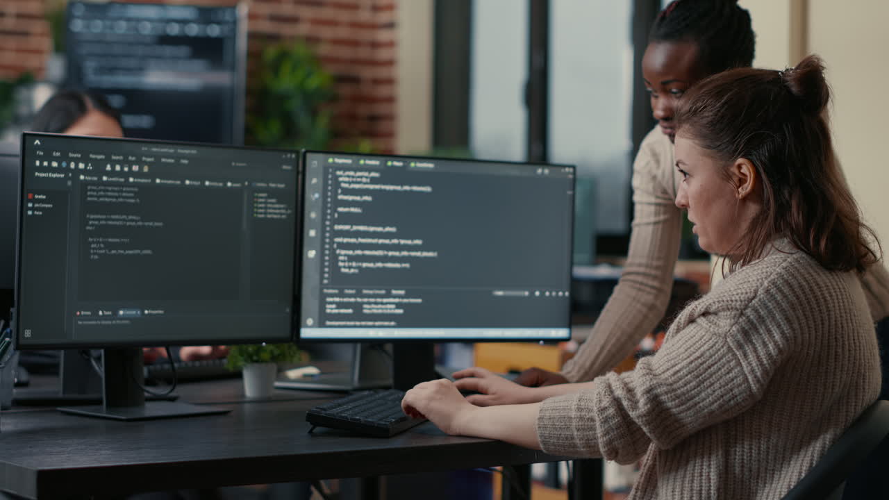 Junior programer writing algorithm under experienced colleague supervision sitting at desk with multiple screens