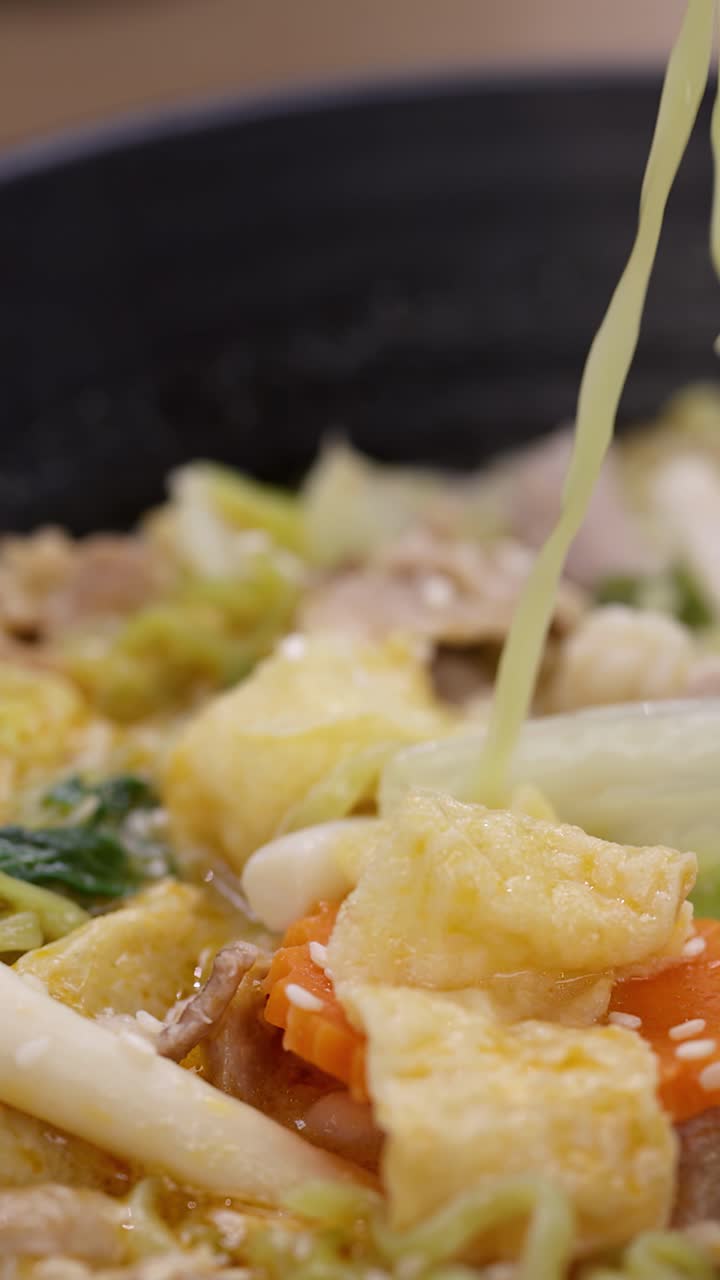 Primer plano de palillos levantando fideos verdes de una sopa humeante de verduras y cerdo con iluminación cálida