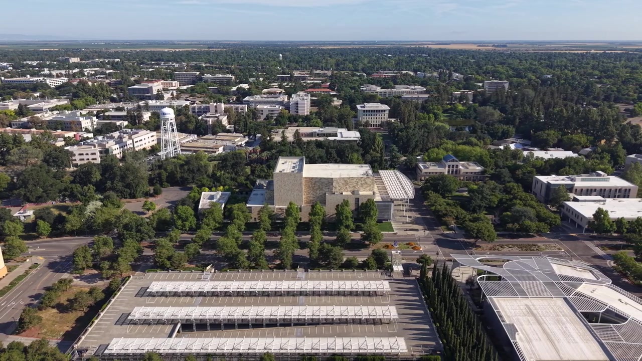 Prospettiva aerea dall'alto in volo sopra il campus dell'Università della California, Davis, che mostra architettura moderna, strutture di parcheggio con pannelli solari e il paesaggio verdeggiante circostante.