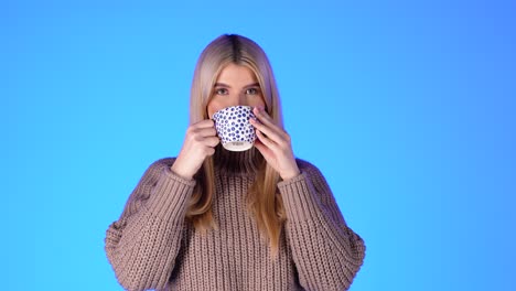 portrait of pretty blonde woman drinking from coffee mug, looking at camera against blue background, studio shot