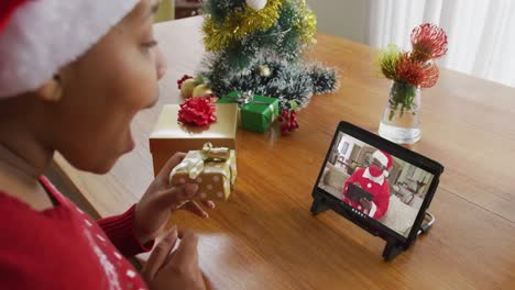 Mujer-Afroamericana-Con-Gorro-De-Papá-Noel-Usando-Tableta-Para-Videollamada-Navideña,-Con-Papá-Noel-En-Pantalla