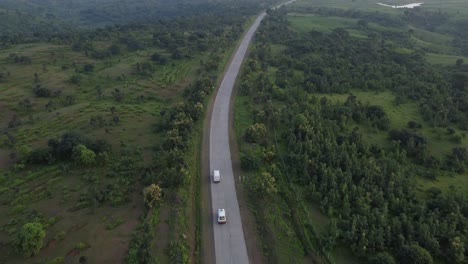 Drone-Siguiendo-A-Una-Ambulancia-Que-Adelanta-A-Una-Furgoneta-De-Transporte-De-Mercancías-Que-Viaja-Por-Una-Carretera-India-Rural-Remota-De-Buena-Calidad-Con-Los-Alrededores-De-Bosques-Naturales-Y-Tierras-De-Cultivo-Verdes-Y-Casas-De-Aldea