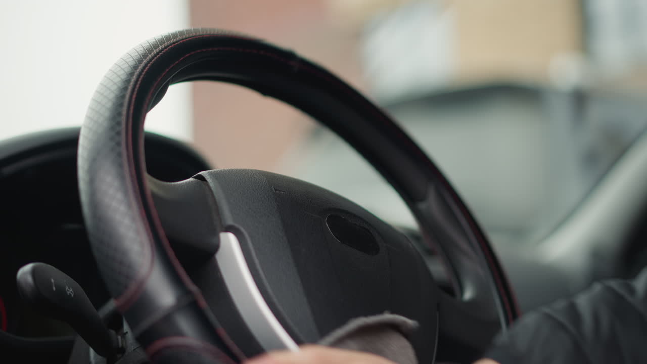 Hands of commuter wiping steering wheel with towel in circular motion inside modern car interior showing quilted seat cover and blurred dashboard background under bright natural daylight