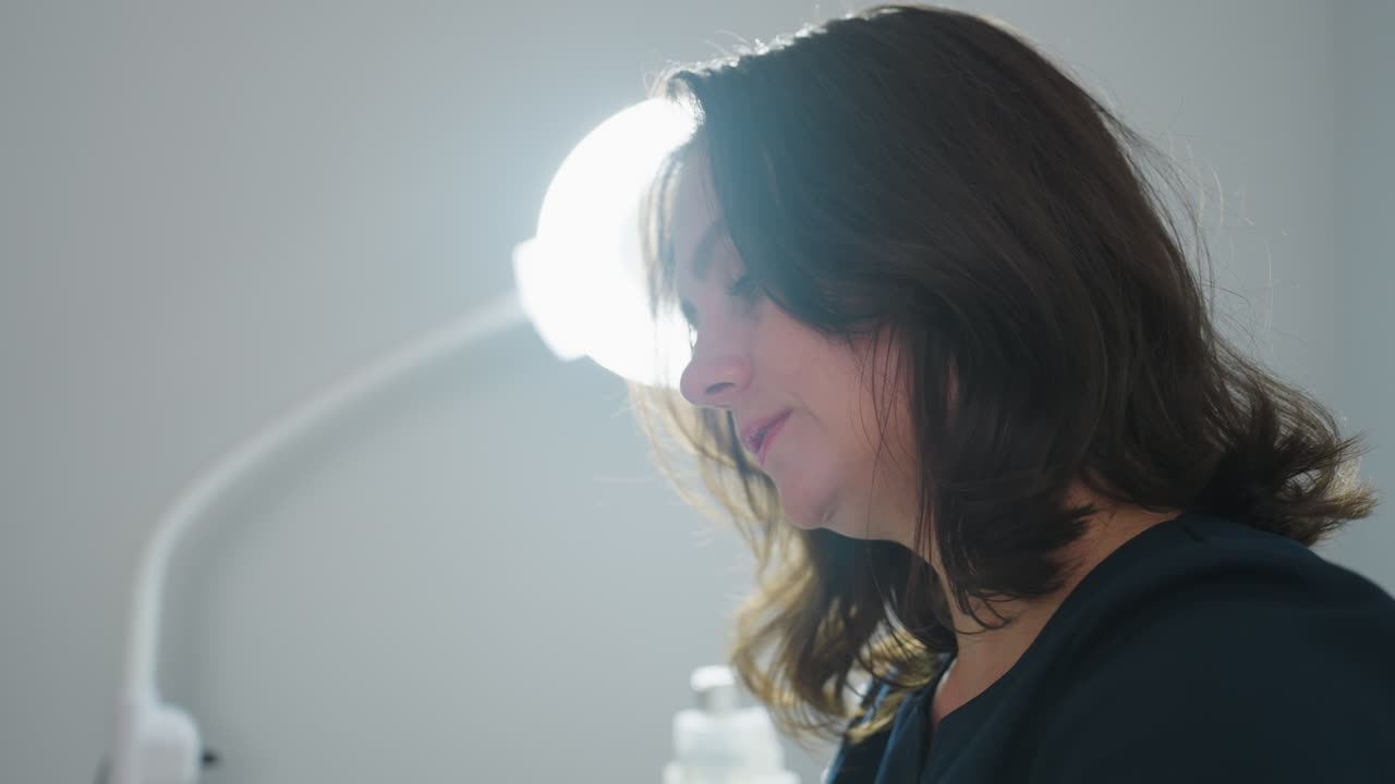 Portrait of cosmetologist near ring light moving head sideways with focused expression during patient session in spa clinic under bright illumination conveying professional care and wellness support