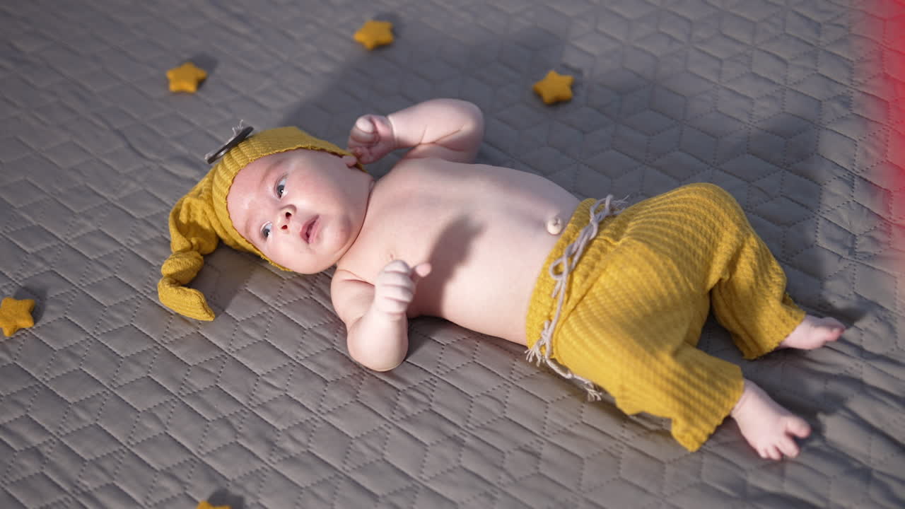 Adorable child in yellow hat and pants lying on the bed face up. Baby in yellow costume with decorative toy stars. View from above.