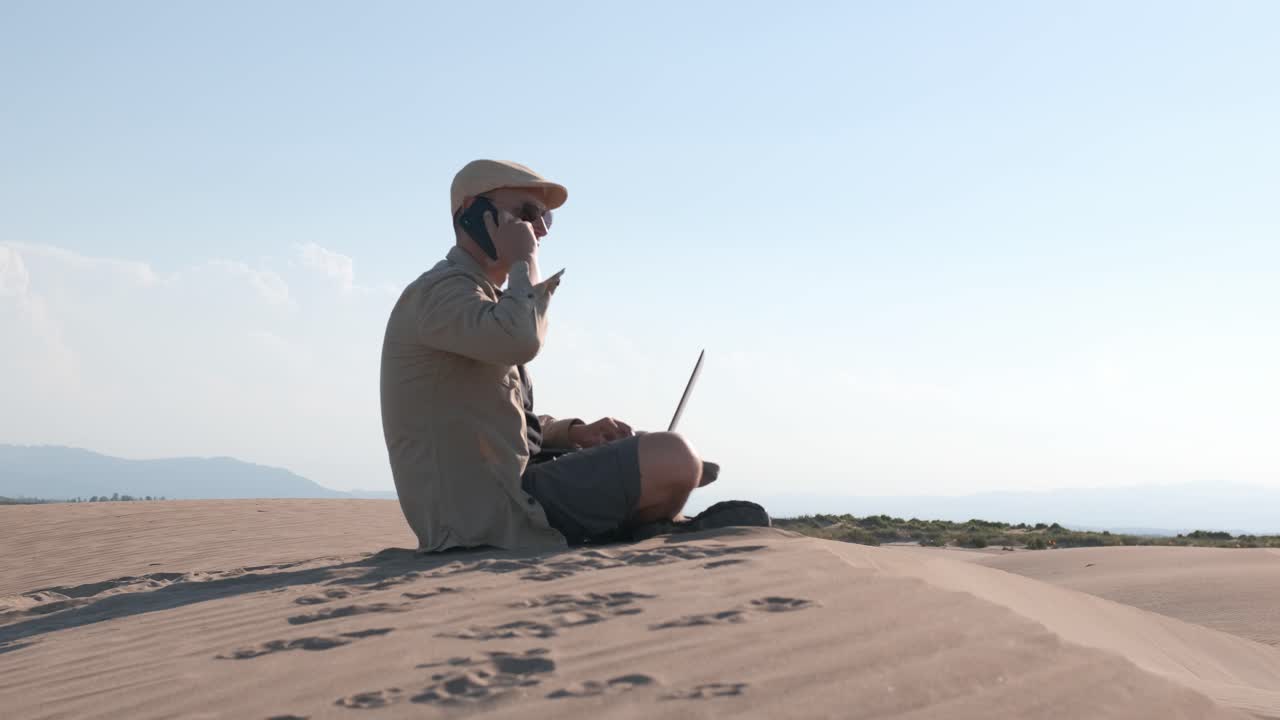 Male traveler sitting on sandy dune using laptop. Digital nomad concept