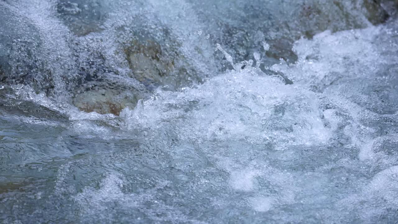 Clear, turbulent river water rushes over rocks in daylight, captured with a static camera. Natural lighting highlights the dynamic, textured surface and movement