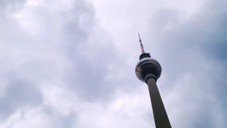 as dawn casts a subtle light, the fernsehturm at alexanderplatz reaches skyward, standing firm against a backdrop of grey clouds scurrying across the awakening sky