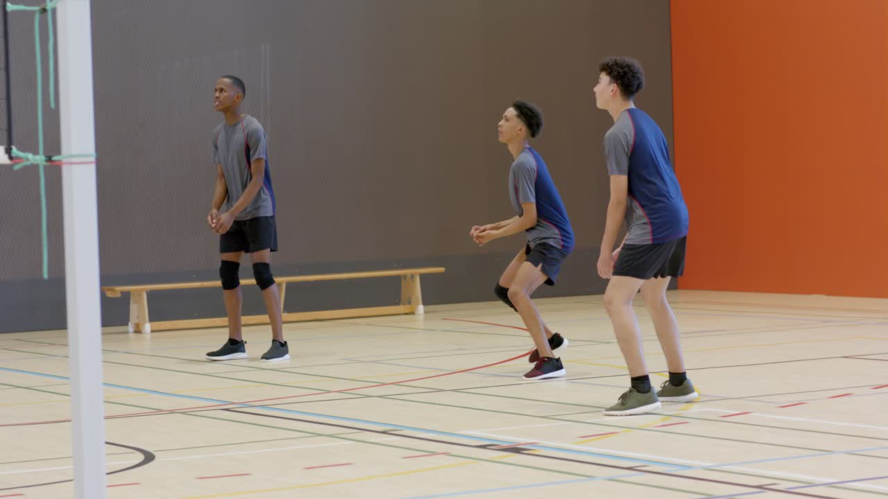 Male volleyball teammates reacting to opponent serve on gym court, digging ball to keep rally going