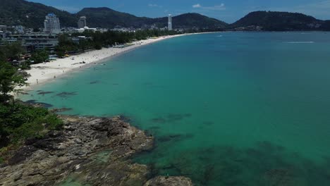 Drone zooms out revealing vibrant blue waters, beach, and urban skyline surrounded by lush hills in Phuket, Thailand