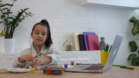 Una-Niña-Pequeña-Y-Seria-Estudia-Escritura-A-Mano-En-Línea-Usando-Una-Computadora-Portátil-En-Casa,-Una-Niña-Pequeña-Y-Feliz-Toma-Una-Lección-O-Clase-En-Internet