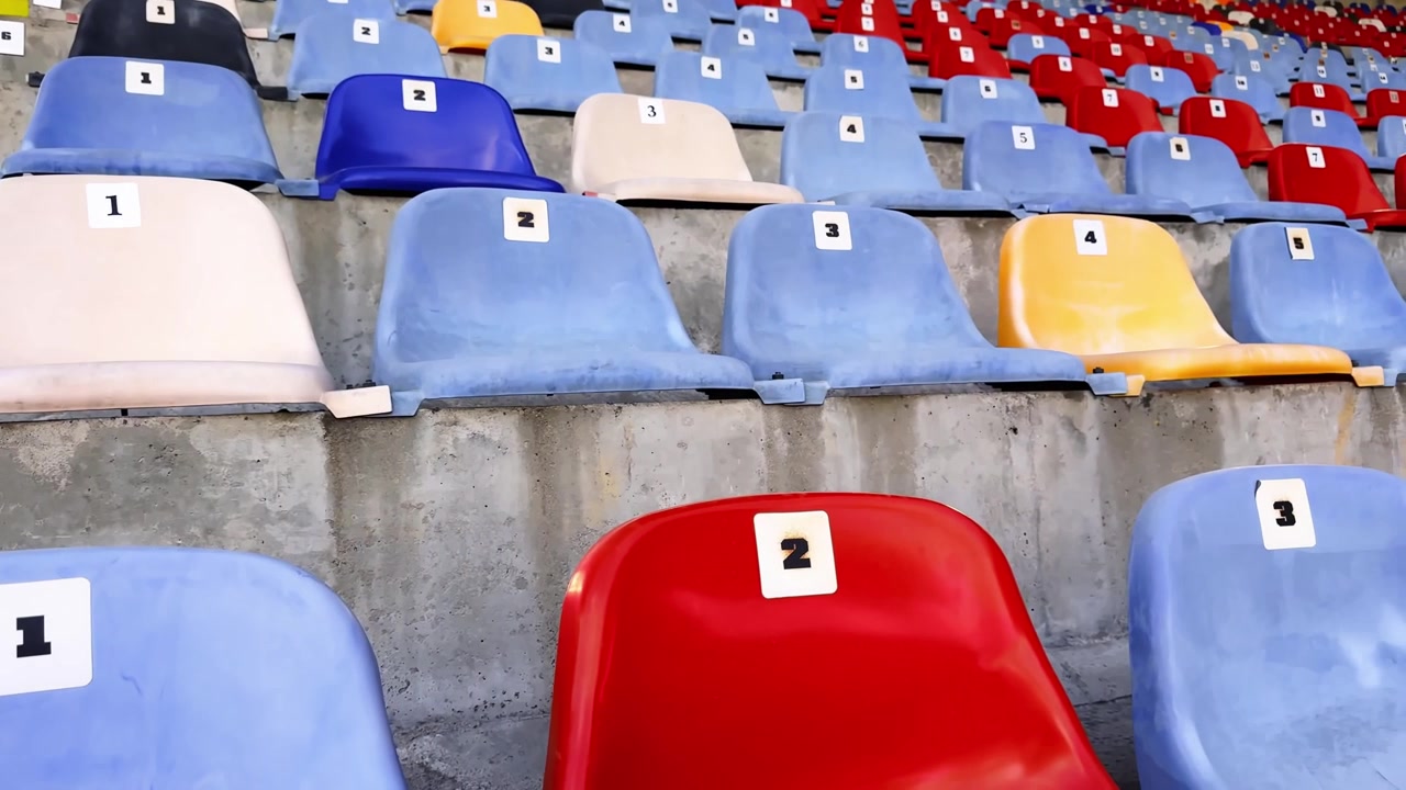 Close-up of numbered plastic stadium seats in red, blue, white and yellow, with concrete rows and bright yellow stairs in an empty sports venue.