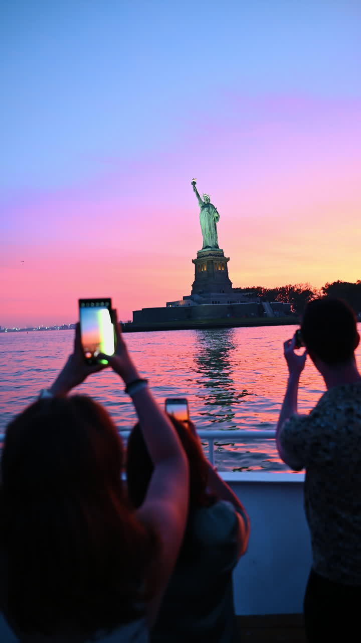 New York, USA, 2 agosto 2025: Turisti fotografano la Statua della Libertà al tramonto dal ponte di un traghetto. Sagome di persone che scattano foto alla Statua della Libertà contro un cielo rosa vibrante al tramonto nel porto di New York.