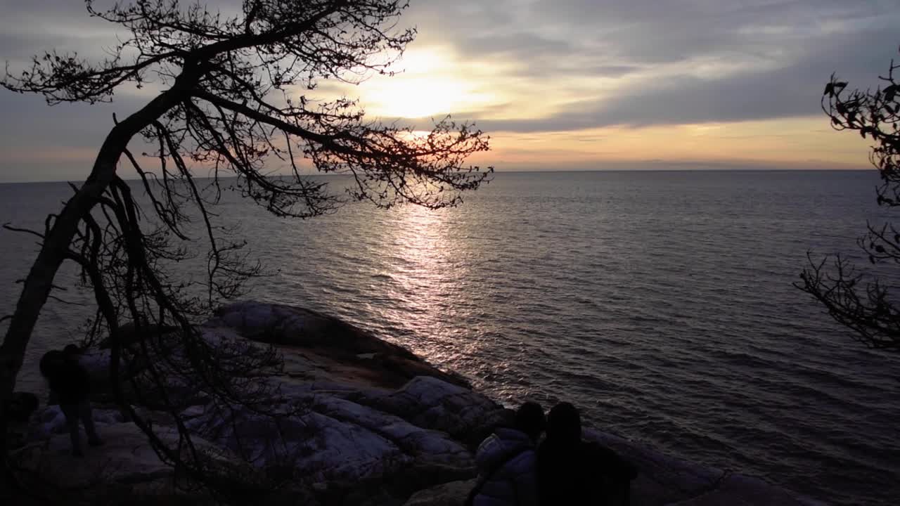 Coppia in silhouette accovacciata a guardare il tramonto da una roccia, ripresa in tilt lunga.