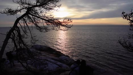 Silhouetted-couple-huddle-watching-sunset-from-rock,-tilt-long-shot