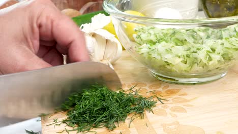 chopping dill on a cutting wooden board. making tzatziki greek or mediterranean sauce or dip for a party. dip or sauce in a green bowl on a wooden cutting board with dill, green napkin and a grater.
