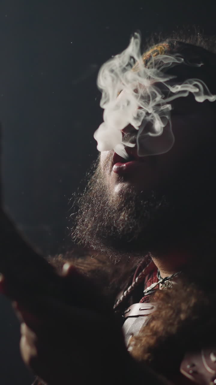 Bearded man in traditional Siberian shaman clothes breathes out smoke clouds on black background closeup. Smoking psychotropic herbs