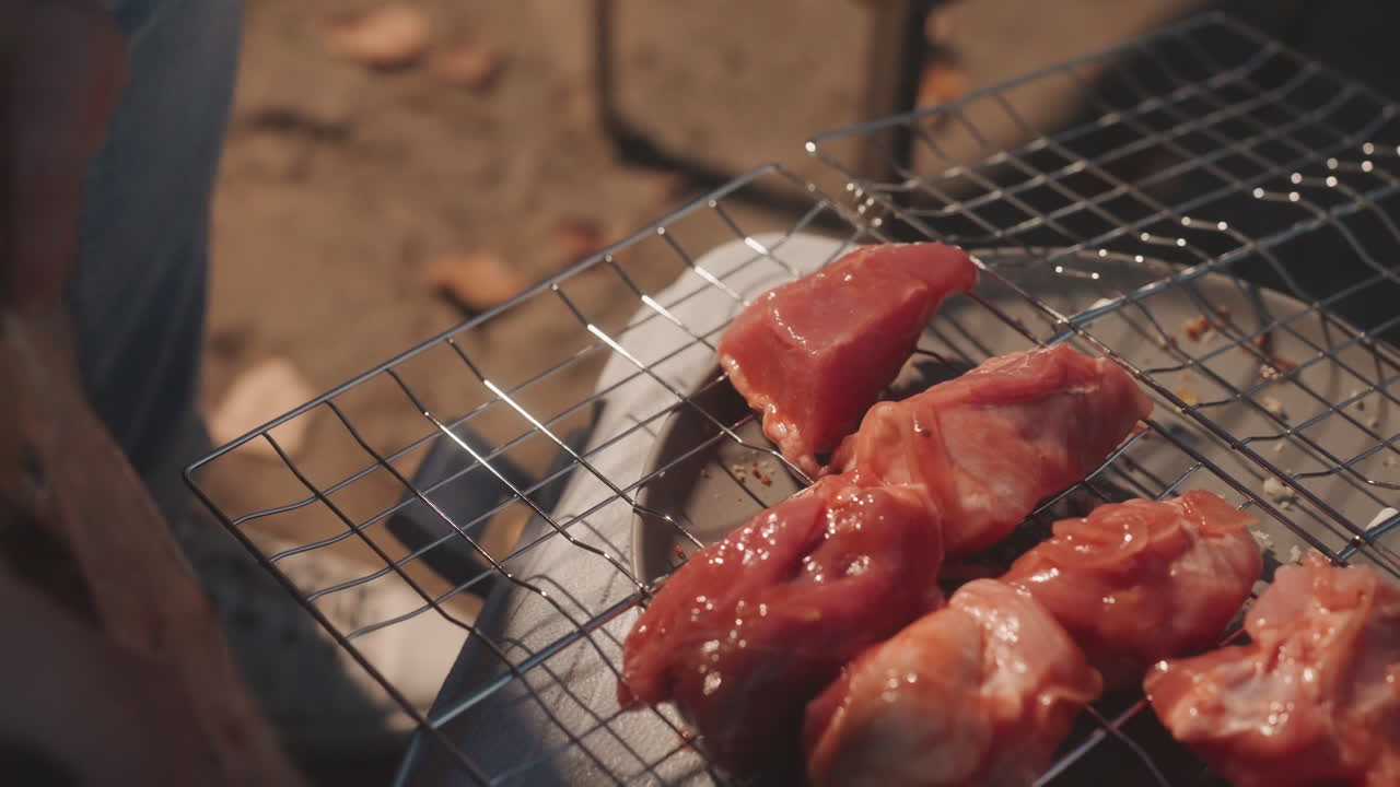 Seorang kemping menata ayam marinasi di atas panggangan, area perkemahan yang diterangi matahari, tangan menekan potongan daging ke rak kawat, panas arang, persiapan barbekyu santai di luar ruangan, suasana piknik komunal, gerakan sentuhan