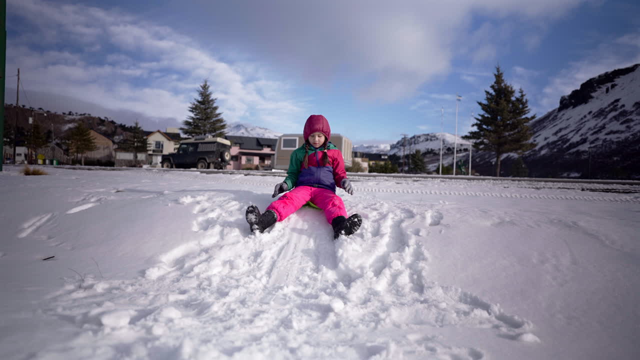Young girl in colorful winter gear have fun dragging a sled through snow, with houses, pine trees, and Andes mountains in the background