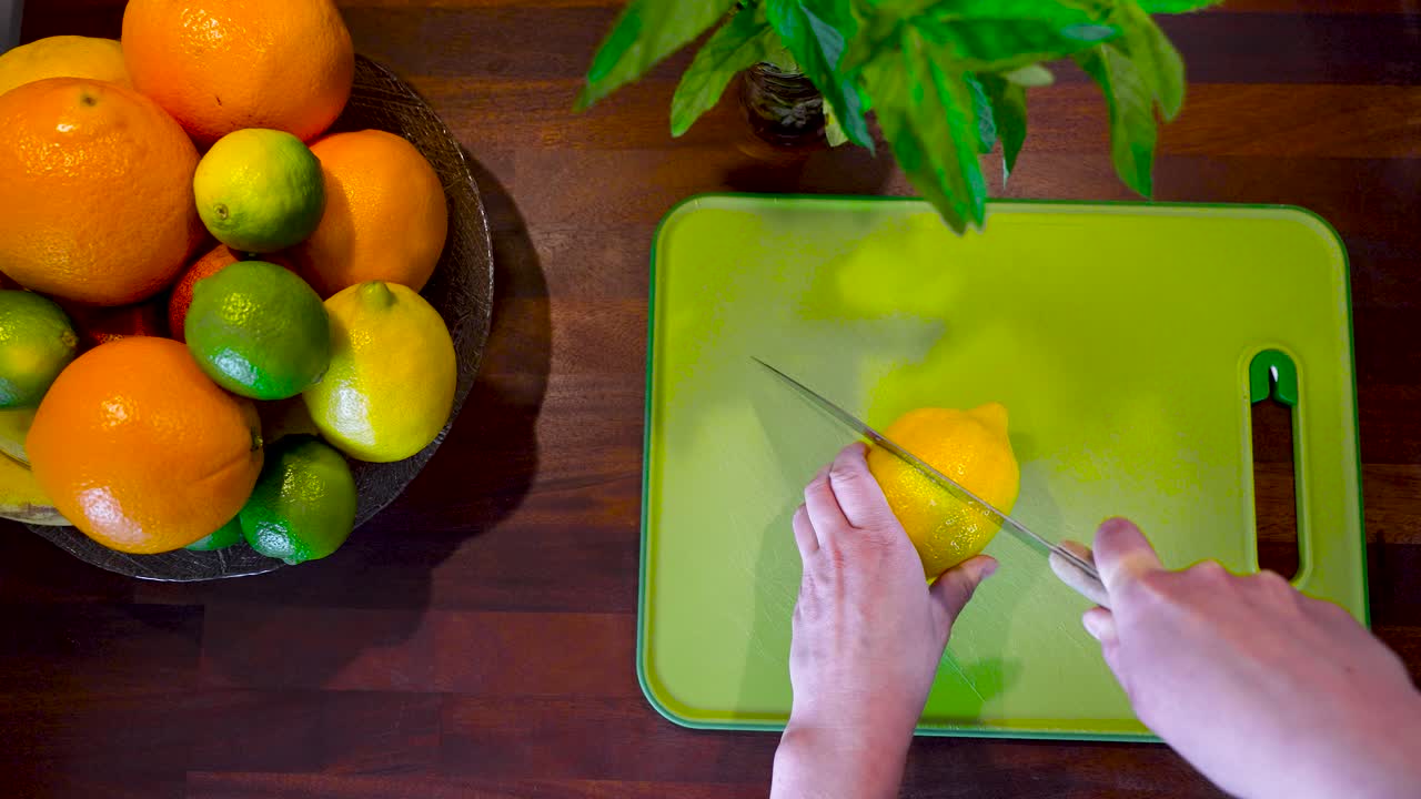 el video muestra un limón amarillo brillante siendo cortado con un cuchillo afilado en una tabla de corte verde que está rodeada de otras frutas cítricas como naranjas, limones, limas y naranjas de sangre