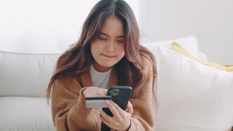 Happy young asian woman holding credit card using instant mobile payments at home. Smiling Female customer shopper making purchase on smartphone receiving cash back concept.
