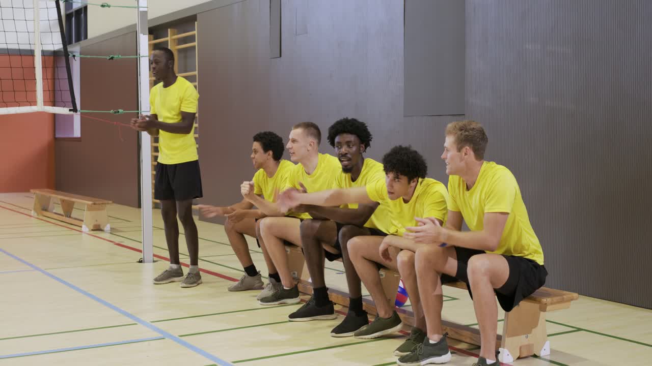 Männliches Volleyballteam in gelben Trikots sitzt auf der Bank in der Turnhalle, reagiert auf Netzspiel, feiert