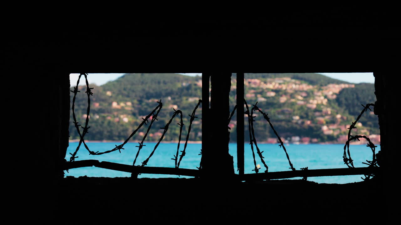 Close up of barbed wire loops along a small window with a blurred view of the sea and a town