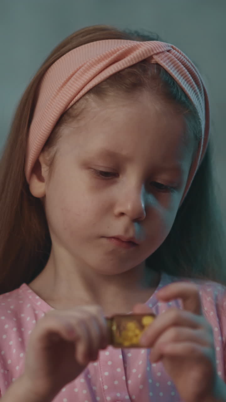 Interested little child with pink headband plays with closed small bottle full of white pills in semi-dark room at home extreme close view slow motion
