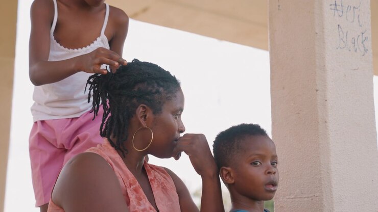 Young girl styles hair of adult woman with braided texture while child sits nearby. scene takes place outdoors under bright daylight styling family bonding
