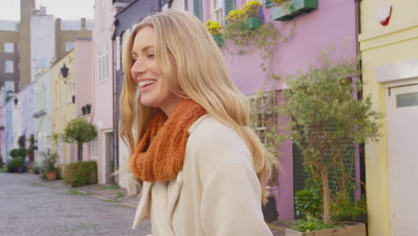 portrait of smiling woman wearing coat and scarf standing on cobbled mews street on visit to city in autumn or winter - shot in slow motion