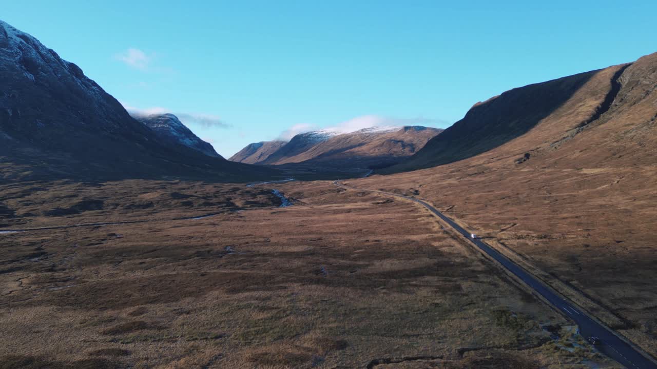 Valle de Glencoe con una carretera sinuosa entre montañas pintorescas, cielo azul claro, vista aérea.