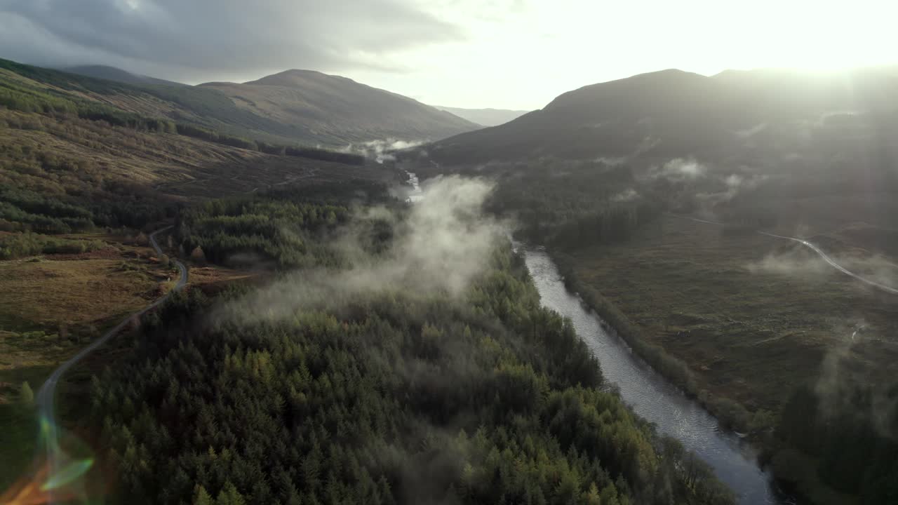 imágenes aéreas de drones que vuelan lentamente hacia atrás por encima de un río, una carretera y un bosque de árboles de coníferas mientras una nube baja abraza las copas de los árboles y el sol se pone detrás de las montañas