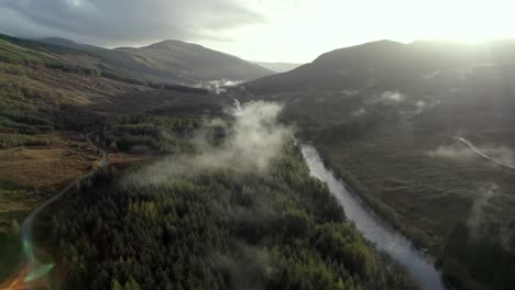 Aerial-drone-footage-flying-slowly-backwards-high-above-a-river,-road-and-forest-of-conifer-trees-while-low-hanging-cloud-hugs-the-treetops-and-the-sun-sets-behind-mountains